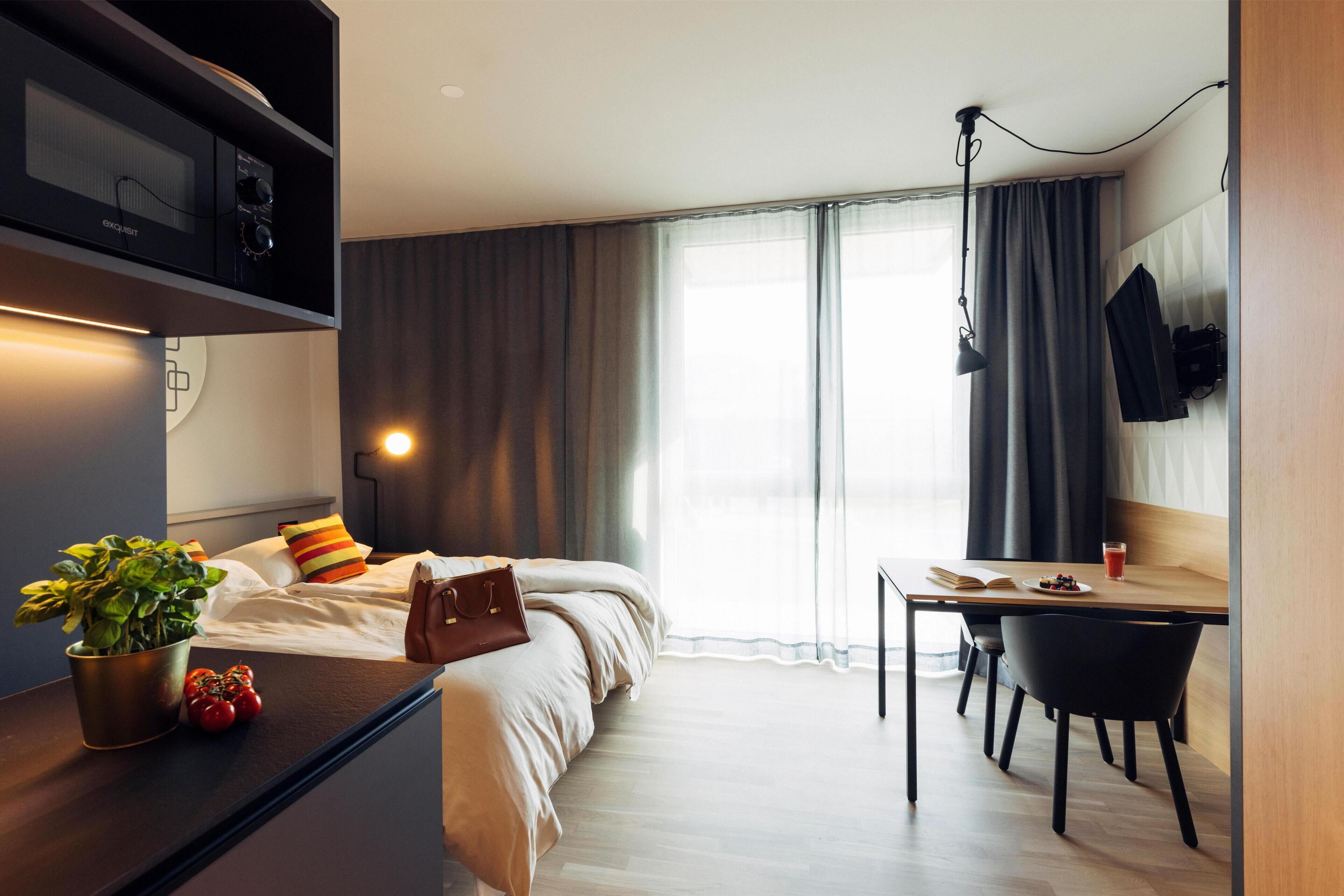 Et moderne hotelværelse med seng, sengebord, skrivebord, stol og et stort vindue med gardiner.