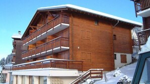 En træchalet med balkon, trappe og en bil foran.