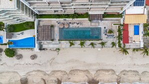 Ovenudblik over en feriepark med en central swimmingpool, flere loungeområder og en strand.