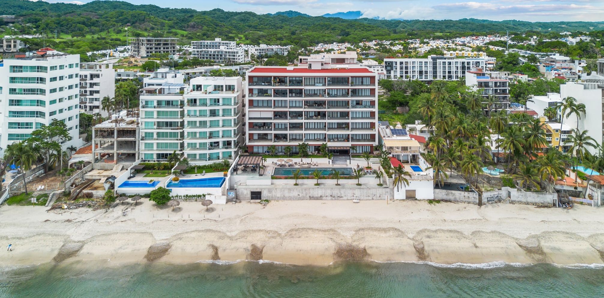 Hotel à beira-mar com vários andares, piscina e praia ao primeiro plano.