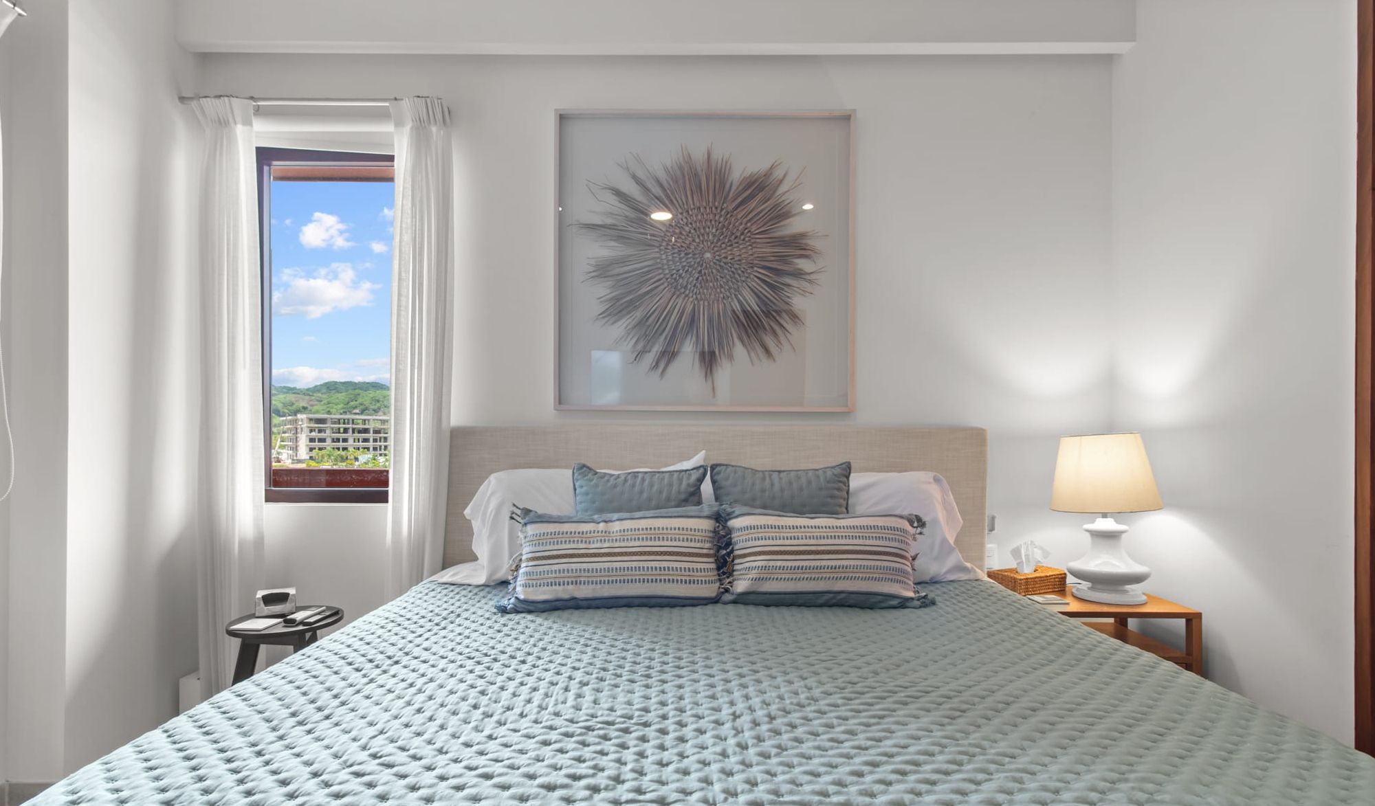 Ein Hotelzimmer mit einem großen Bett, einem gerahmten Kunstwerk darüber, Blick durch das Fenster und einem Nachttisch mit Lampe.