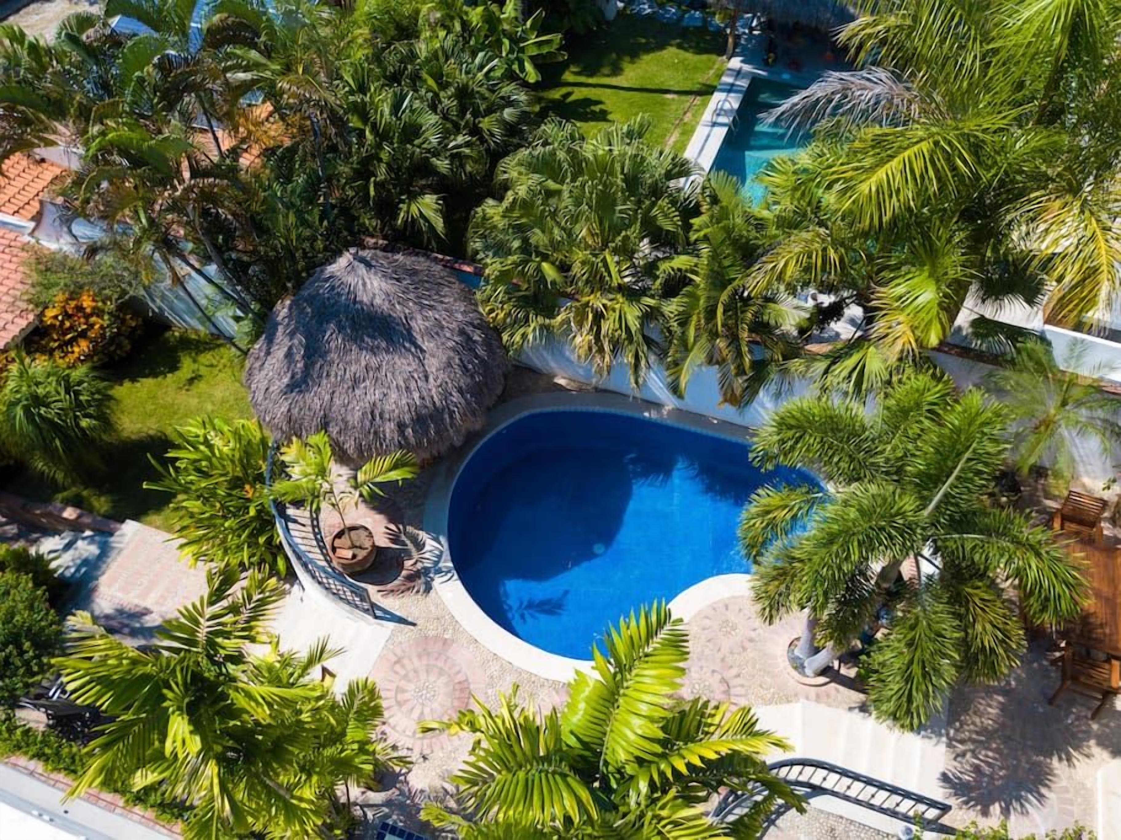 An aerial view of a resort with a central swimming pool surrounded by lush greenery and palm trees.
