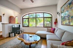 A living room with a large arched window, a sofa, a coffee table, and a TV. - Villa Delfin: Golden Zone 3BR with Private Pool (Bucerías)