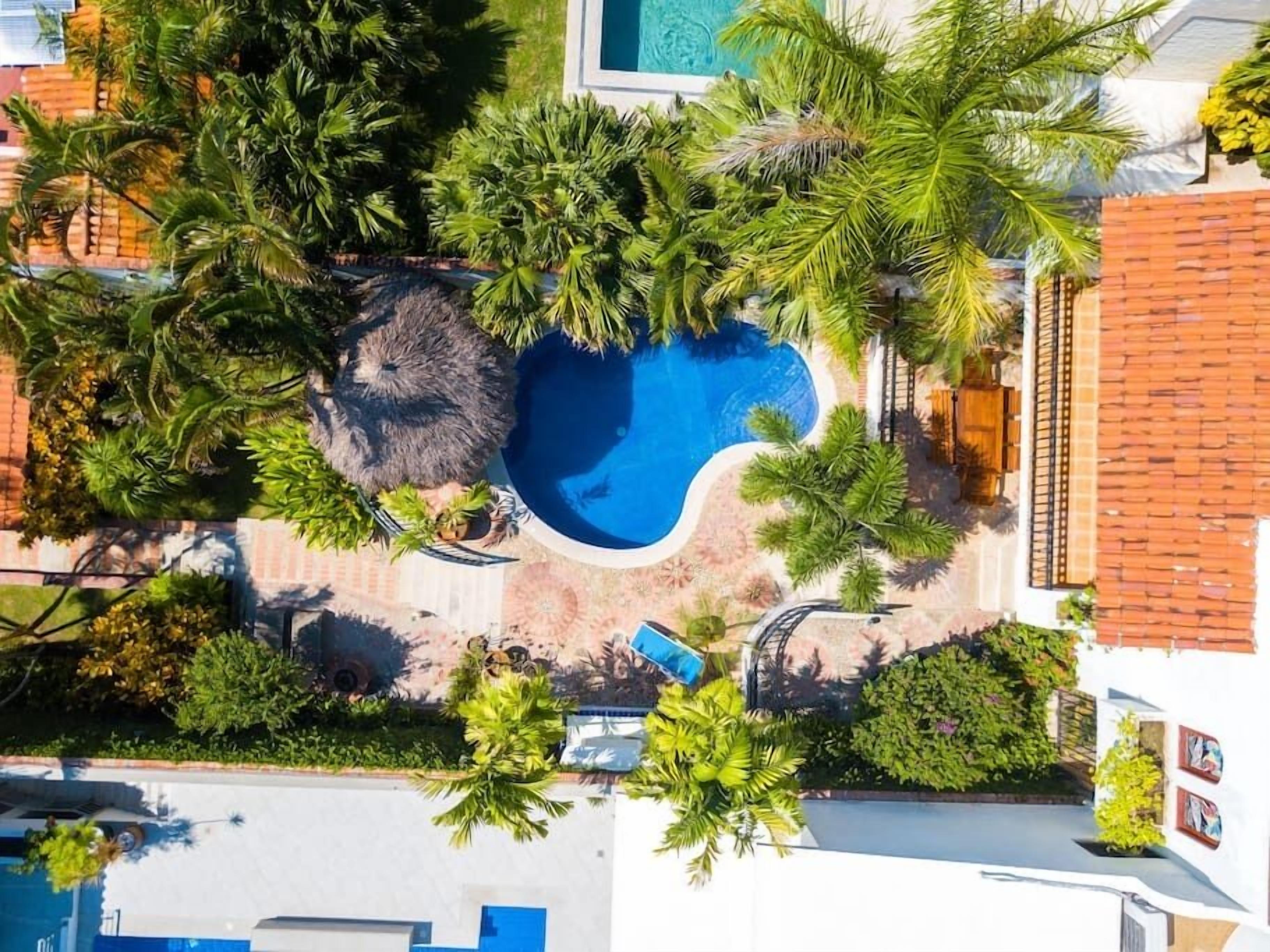 An aerial view of a resort with a central swimming pool surrounded by lush greenery and palm trees.