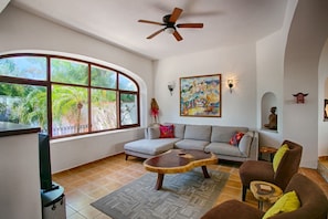 A living room with a large window, a ceiling fan, a sofa, a coffee table, and a painting on the wall. - Villa Delfin: Golden Zone 3BR with Private Pool (Bucerías)