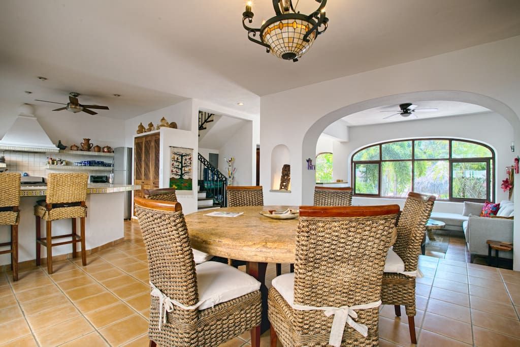 A spacious dining area with a round table and wicker chairs, a kitchen with a ceiling fan, and large windows offering a view of greenery.