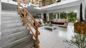 A spacious living area with a modern staircase, comfortable seating, and large windows offering a view of greenery outside.