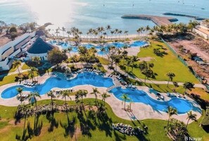 An aerial view of a resort with multiple swimming pools, palm trees, and a beach area.