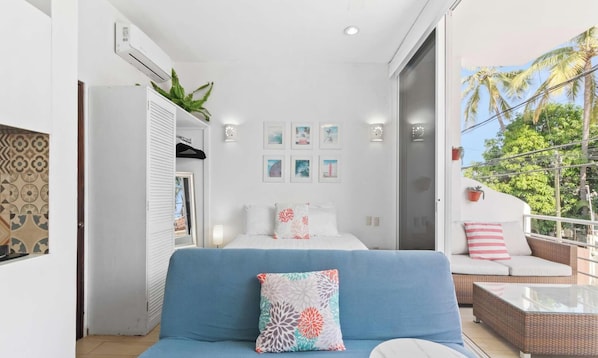 A bedroom with a bed, a blue sofa, a balcony with a view of palm trees, and a door leading to another room. - Golden Zone Studio w Pool - 2 Blocks to Beach (Bucerías)