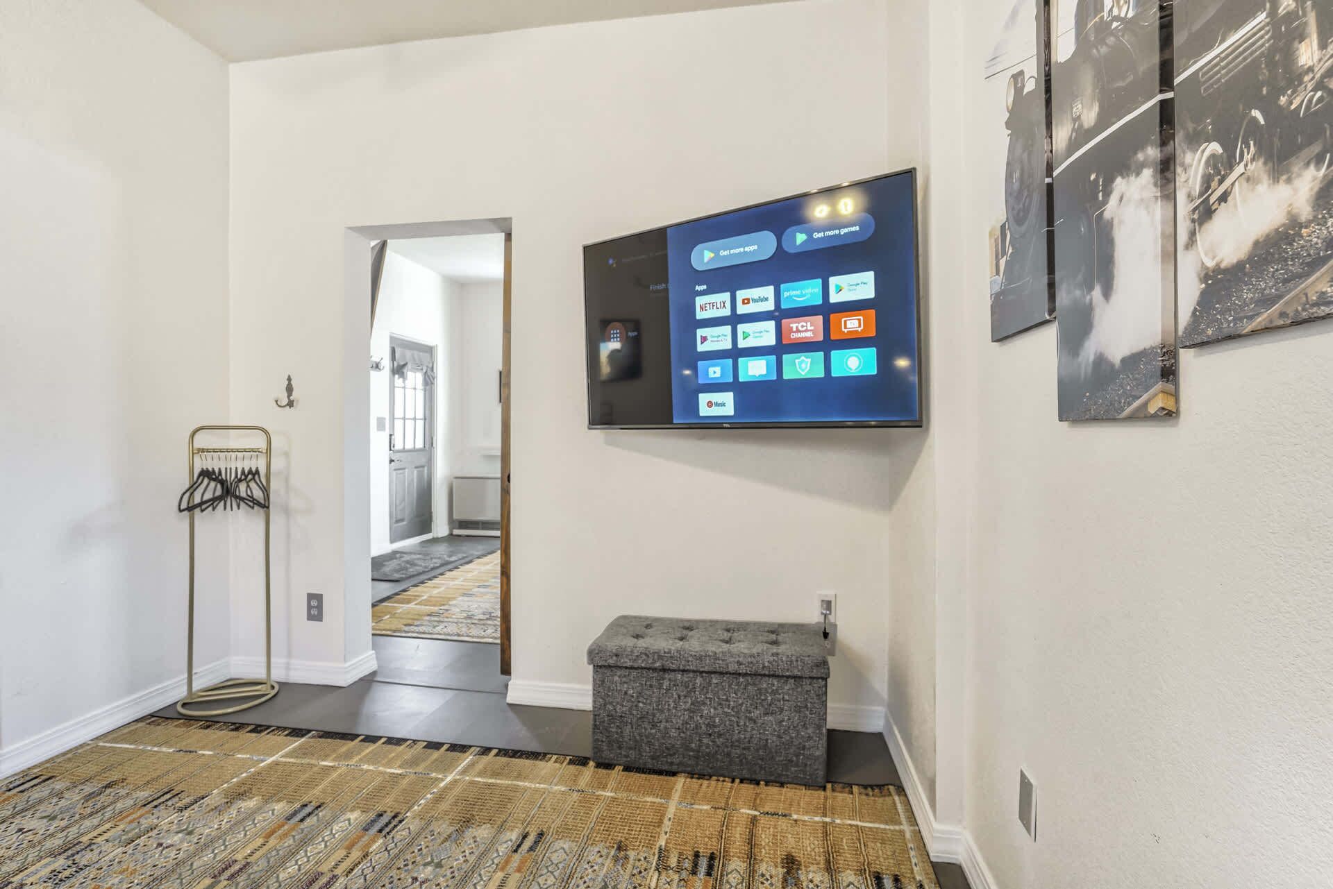 Un couloir avec un téléviseur à écran plat affichant diverses icônes d’applications, un pouf gris, un porte-manteau et un tapis à motifs.