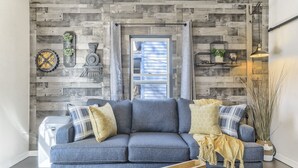 Un salon avec un canapé gris, des coussins décoratifs, une table basse en bois et une fenêtre avec des stores.