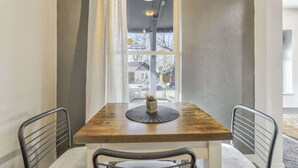 Un coin repas avec une table en bois, deux chaises, un vase de fleurs et une vue par une fenêtre agrémentée d’une grille en fer décorative.