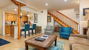 A spacious living area with a wooden coffee table, a sofa, and a blue armchair. A kitchen with wooden cabinets and a dining area with chairs are visible in the background. - Fort Marcy 26 | Walk to Plaza | Kiva Fireplace | Full Kitchen | Hot Tub | Pool | 2 Levels (Santa Fe)