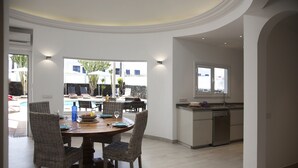 Un coin repas avec des chaises en osier et une table en bois, une cuisine équipée d’un lave-vaisselle, et une vue sur une piscine et un espace de détente extérieur.