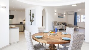 Un espace de vie moderne comprenant un coin repas avec une table en bois et des chaises en osier, une cuisine dotée d’un téléviseur à écran plat et un salon avec un canapé et une grande décoration en forme de cactus.