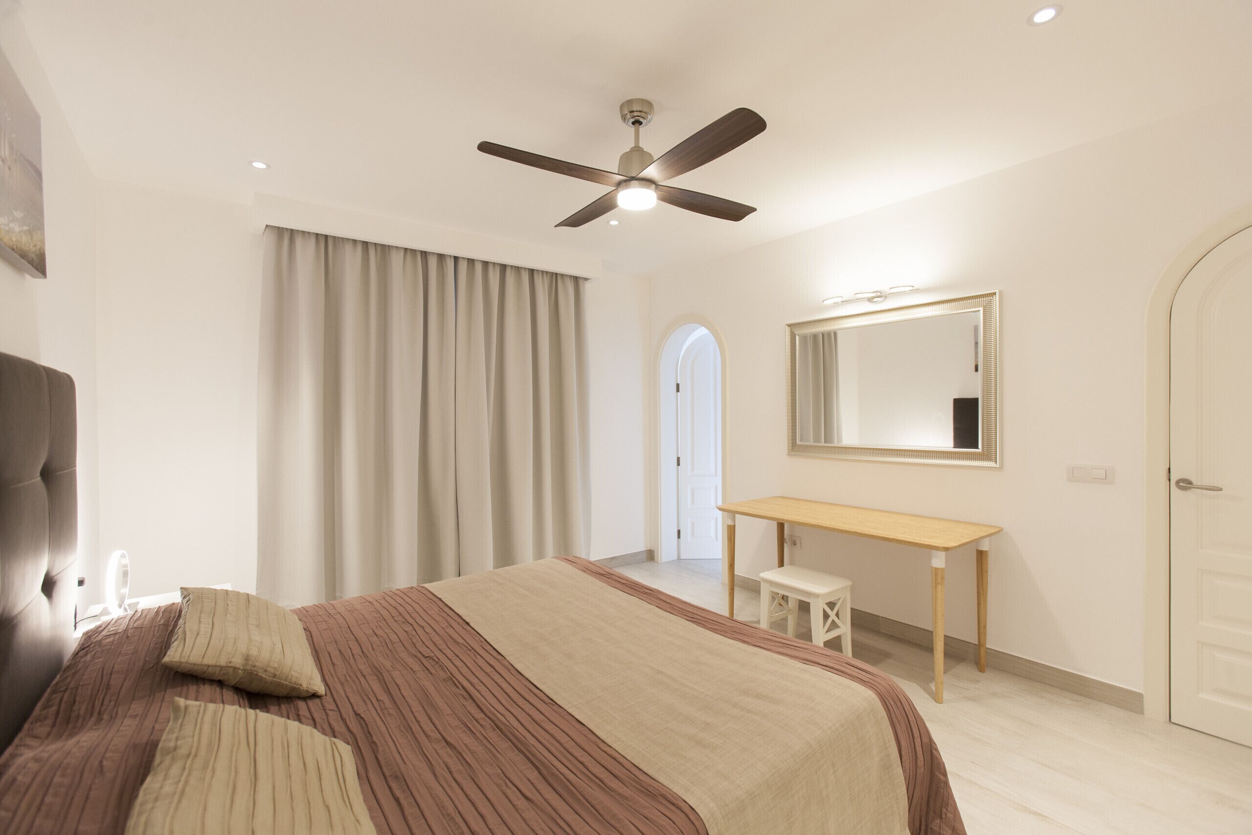 Une chambre à coucher avec un lit, un ventilateur de plafond, une table de chevet, un miroir et une porte.