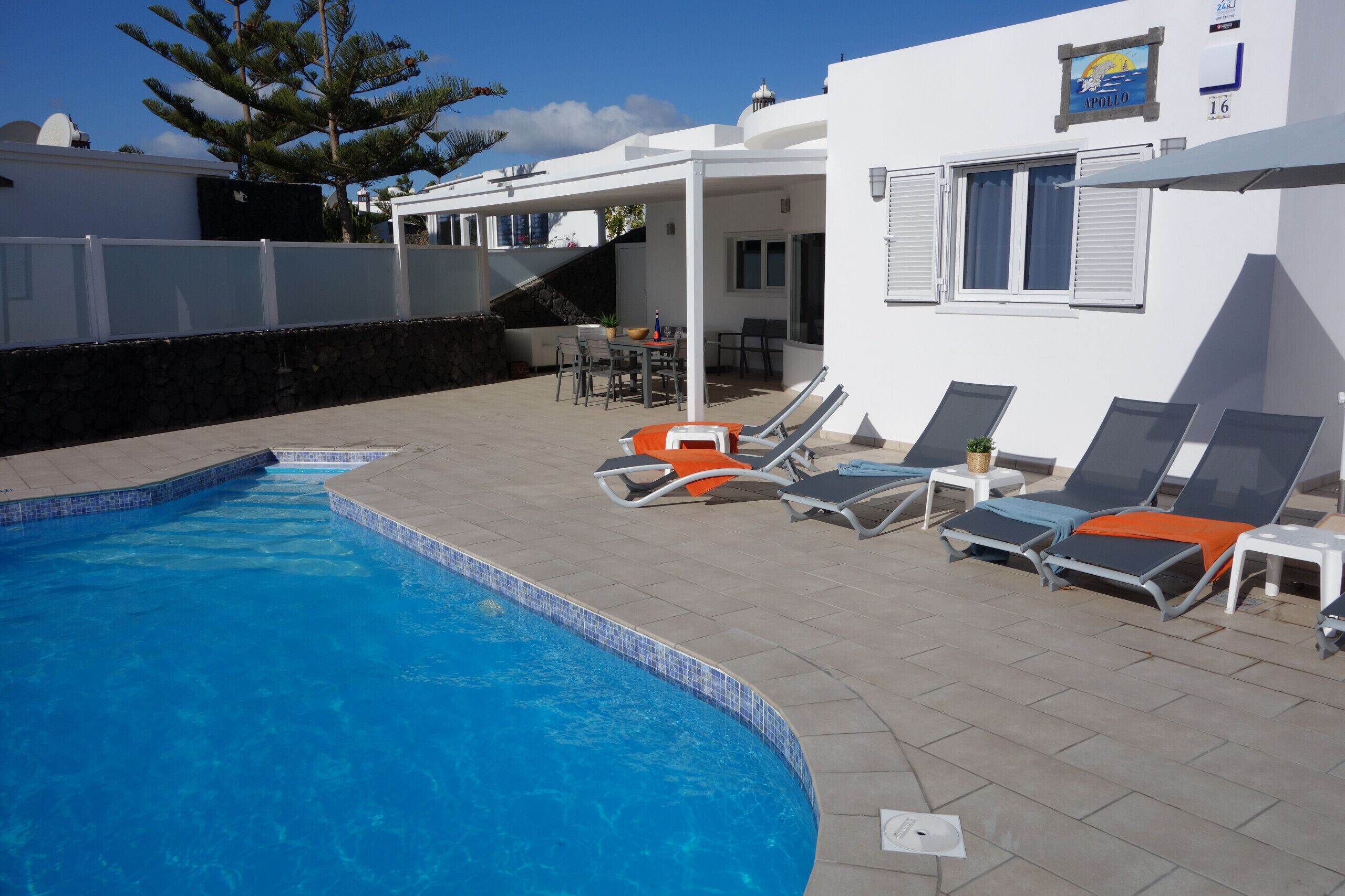 A swimming pool with lounge chairs and a table, surrounded by a white building with a covered patio.
