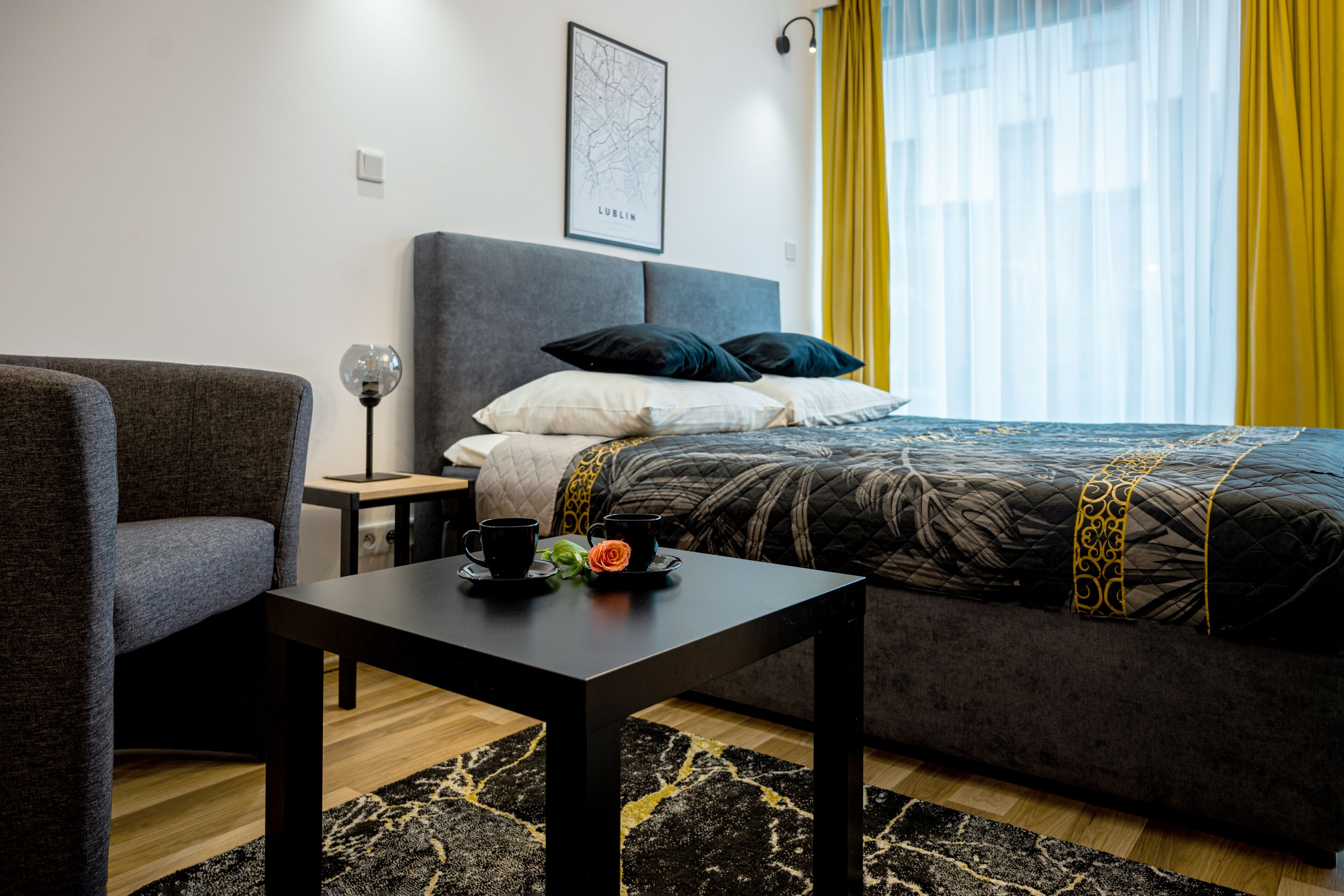 Quarto de hotel moderno com uma cama, uma poltrona cinza, uma mesinha com uma chávena e uma rosa, e um mapa na parede.