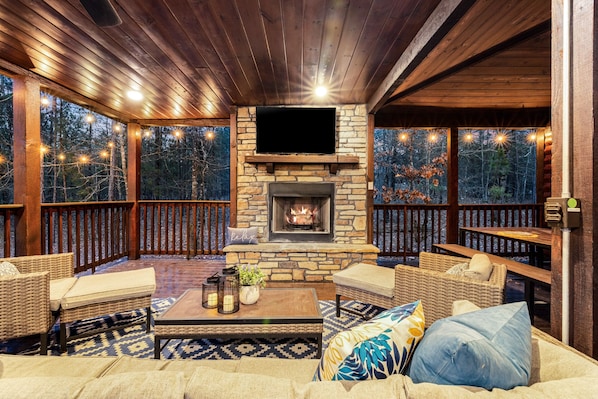 A cozy indoor seating area with wicker furniture, a stone fireplace, and a large window overlooking a forest.