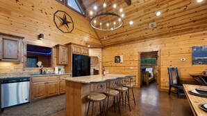 A spacious kitchen with wooden walls, a central island with bar stools, a black refrigerator, and a large window. - 5Kings | Pool Table, Shuffleboard, Playset, Putt Putt | 2min to New Casino! (Broken Bow)