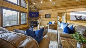 A cozy log cabin interior with a stone fireplace, wooden walls, and a large window. - 5Kings | Pool Table, Shuffleboard, Playset, Putt Putt | 2min to New Casino! (Broken Bow)