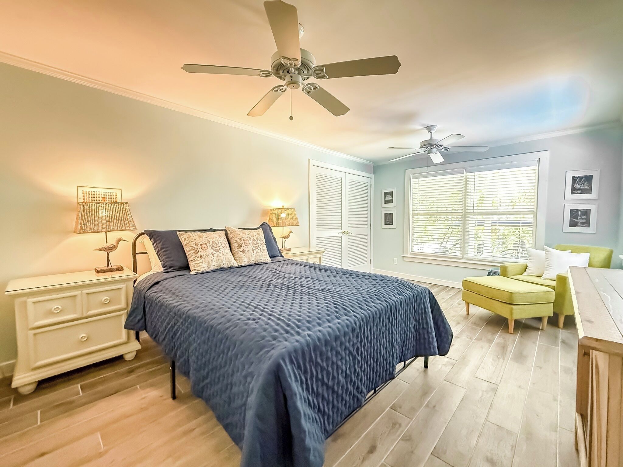 A bedroom with a bed, bedside tables, a desk, a chair, a ceiling fan, and a window with blinds.