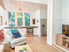 A modern kitchen with white cabinetry, a stainless steel refrigerator, and a built-in oven. There is a dining area with a white sofa and a coffee table. - BLUE MANGO: Tropical Seclusion Steps from the Beach (Captiva)