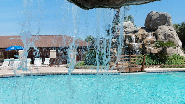 Ein klares, blaues Schwimmbecken mit Wasserfall, Liegestühlen und einem Gebäude mit einem Ziegeldach im Hintergrund.