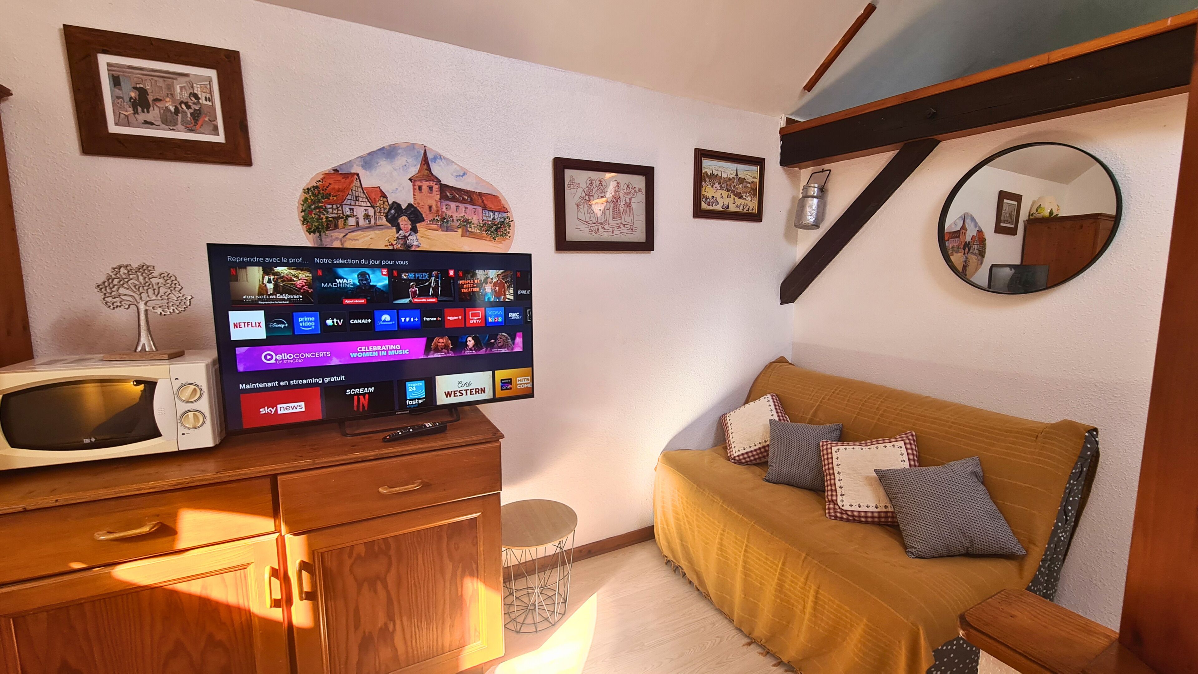 Ein gemütliches Zimmer mit Fernseher, der verschiedene Streaming-Dienste anbietet, einer Holzkommode, einer Couch mit Kissen und gerahmten Bildern an der Wand.