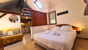 Une chambre à coucher comprenant un lit, une table de chevet, une commode, un miroir, une fenêtre avec des rideaux, un puits de lumière, une petite kitchenette et un plafond en bois.