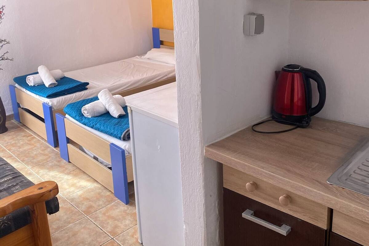 Una habitación pequeña y funcional con dos camas individuales, un escritorio de madera, una tetera roja y un armario pequeño.