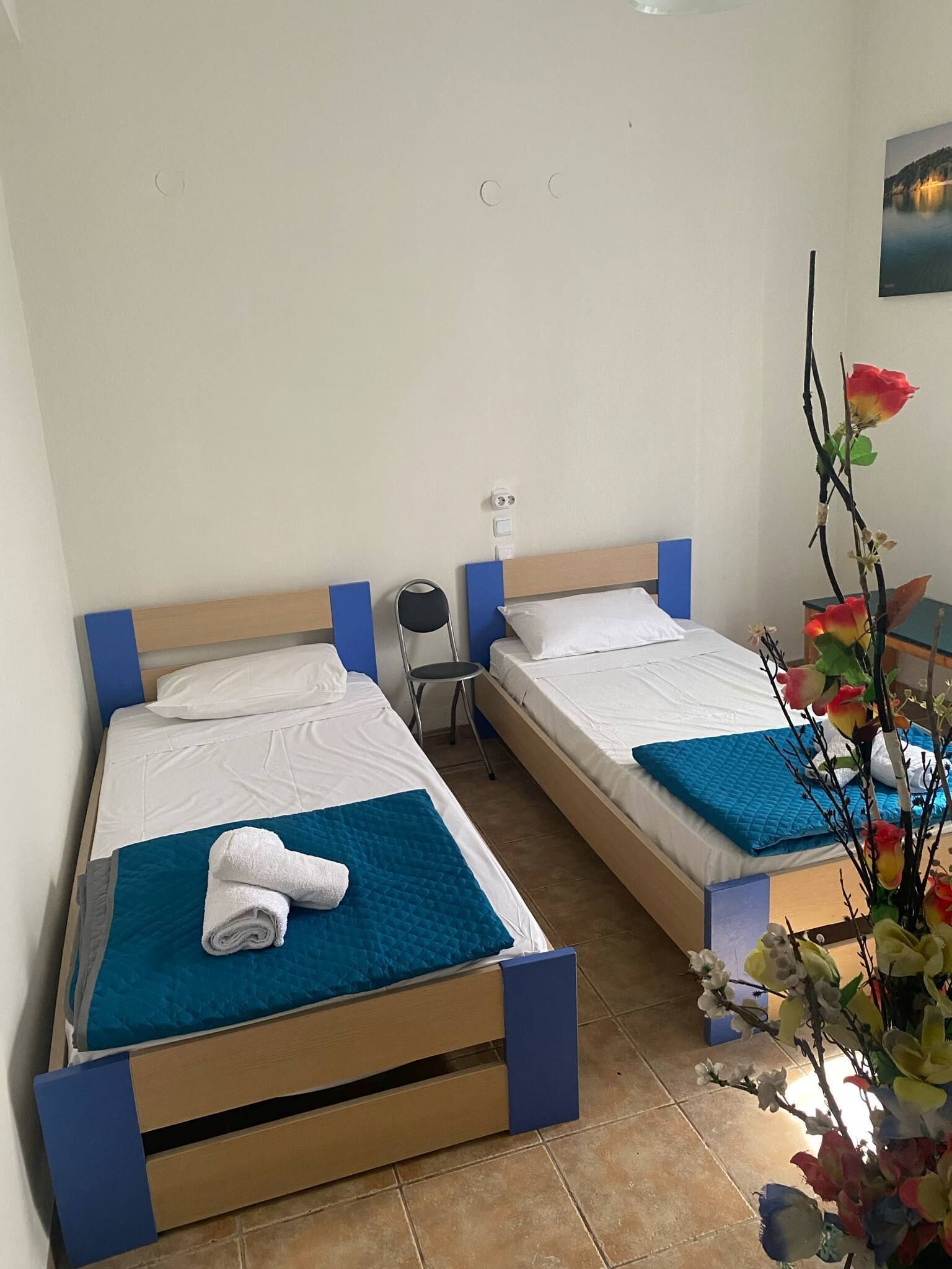 A hotel room with two single beds, each with a blue headboard and white bedding. There is a small table with a chair, a wall-mounted lamp, and a vase with flowers.