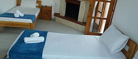 Quarto com três camas individuais, cada uma com uma toalha azul e roupa de cama branca. Há uma mesa de cabeceira de madeira com abajur, lareira e janela com moldura de madeira.