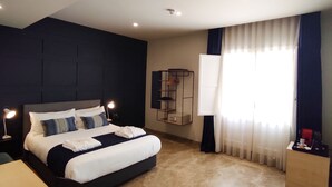 A modern hotel room with a large bed, a desk, a lamp, a shelf, and a window with curtains. - The Manoel Hotel (Valletta)