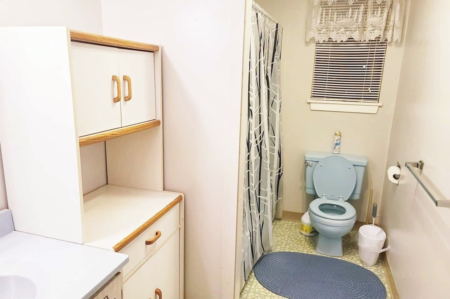 A compact bathroom with a toilet, a sink, a cabinet with drawers, a shower curtain, and a window with blinds.