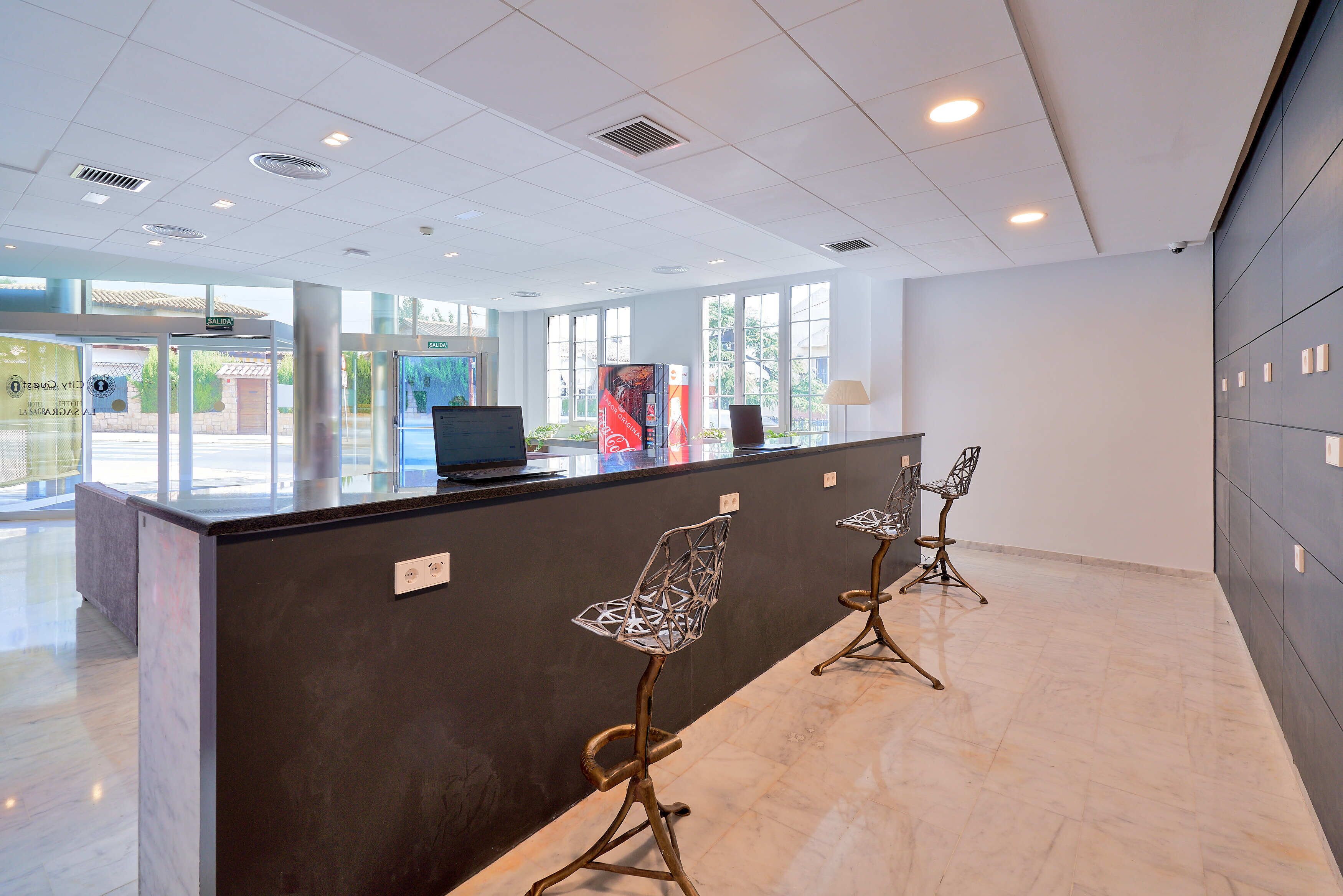A reception area with a dark counter, three metal chairs, a laptop, and a view of the outdoors through large windows.