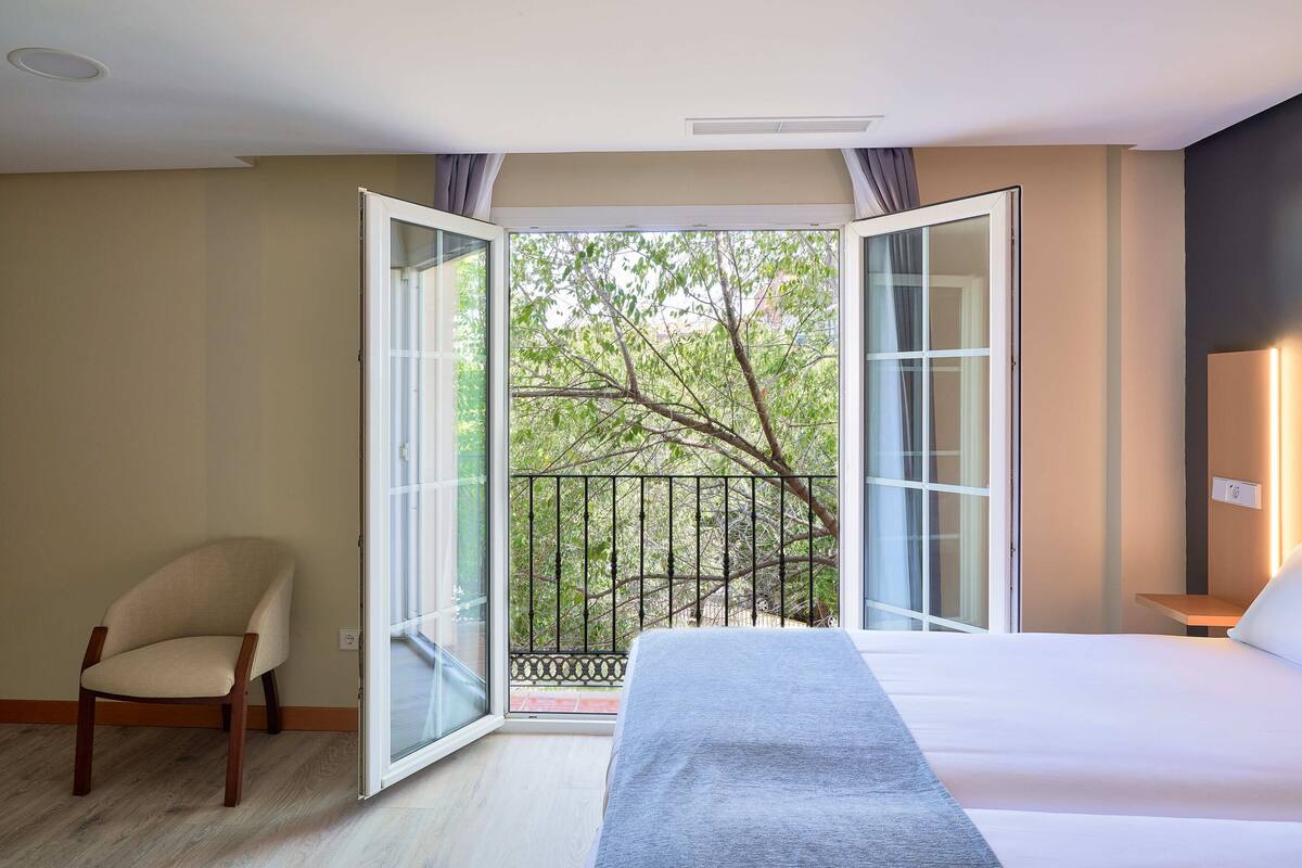 A hotel room with a bed, a chair, and a balcony with a view of trees.