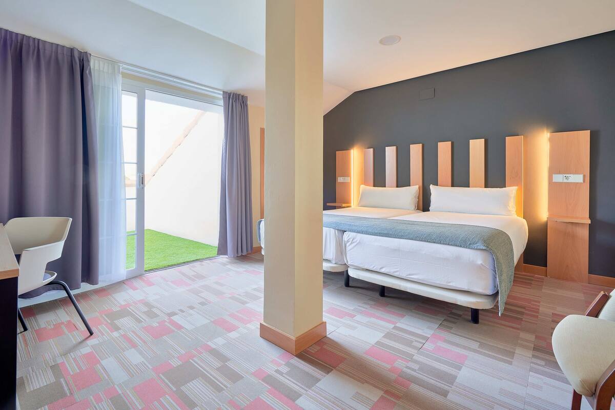 A hotel room with a bed, a desk, and a chair. There is a large window with curtains.