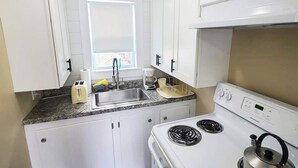 A compact kitchen with white cabinets, a stainless steel sink, a stove, and a kettle.