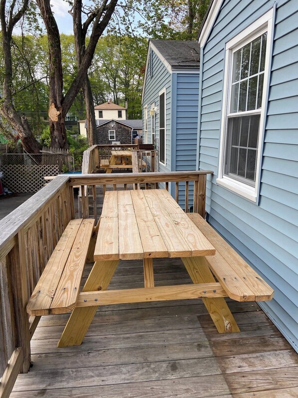 A wooden deck with picnic tables, a blue house, and trees in the background.