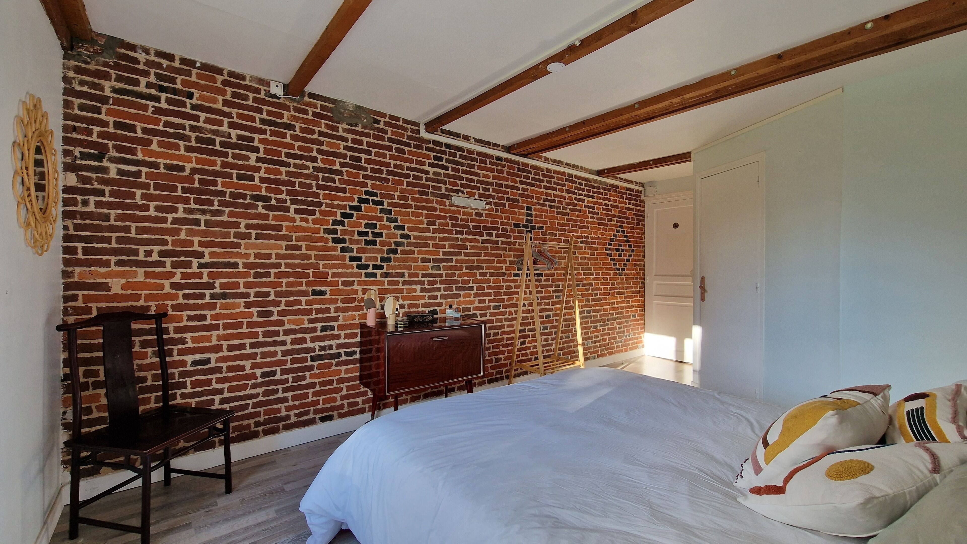 A bedroom with a brick wall, wooden beams, a bed with white bedding, and a wooden chair.