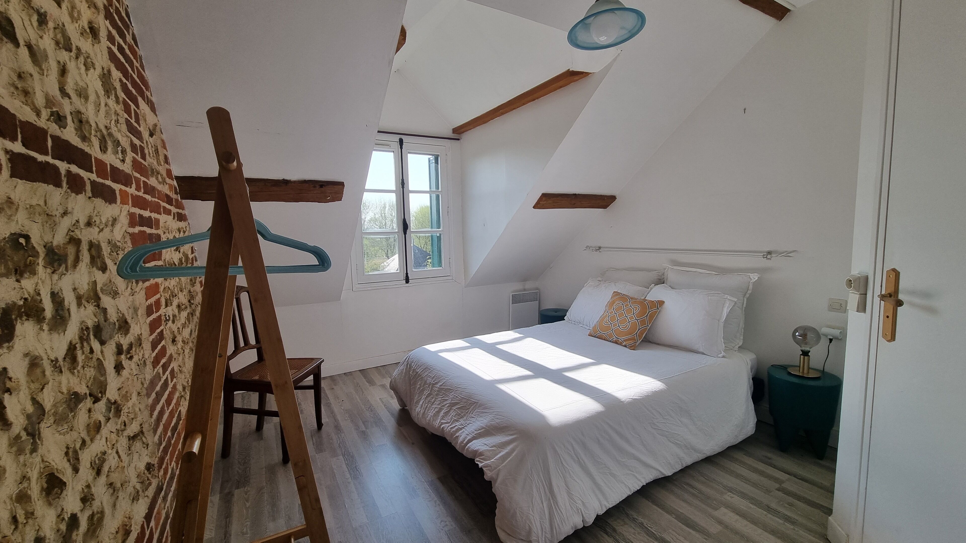Une chambre avec un plafond en pente, un lit avec une literie blanche, une chaise en bois, un luminaire suspendu et un mur en briques.