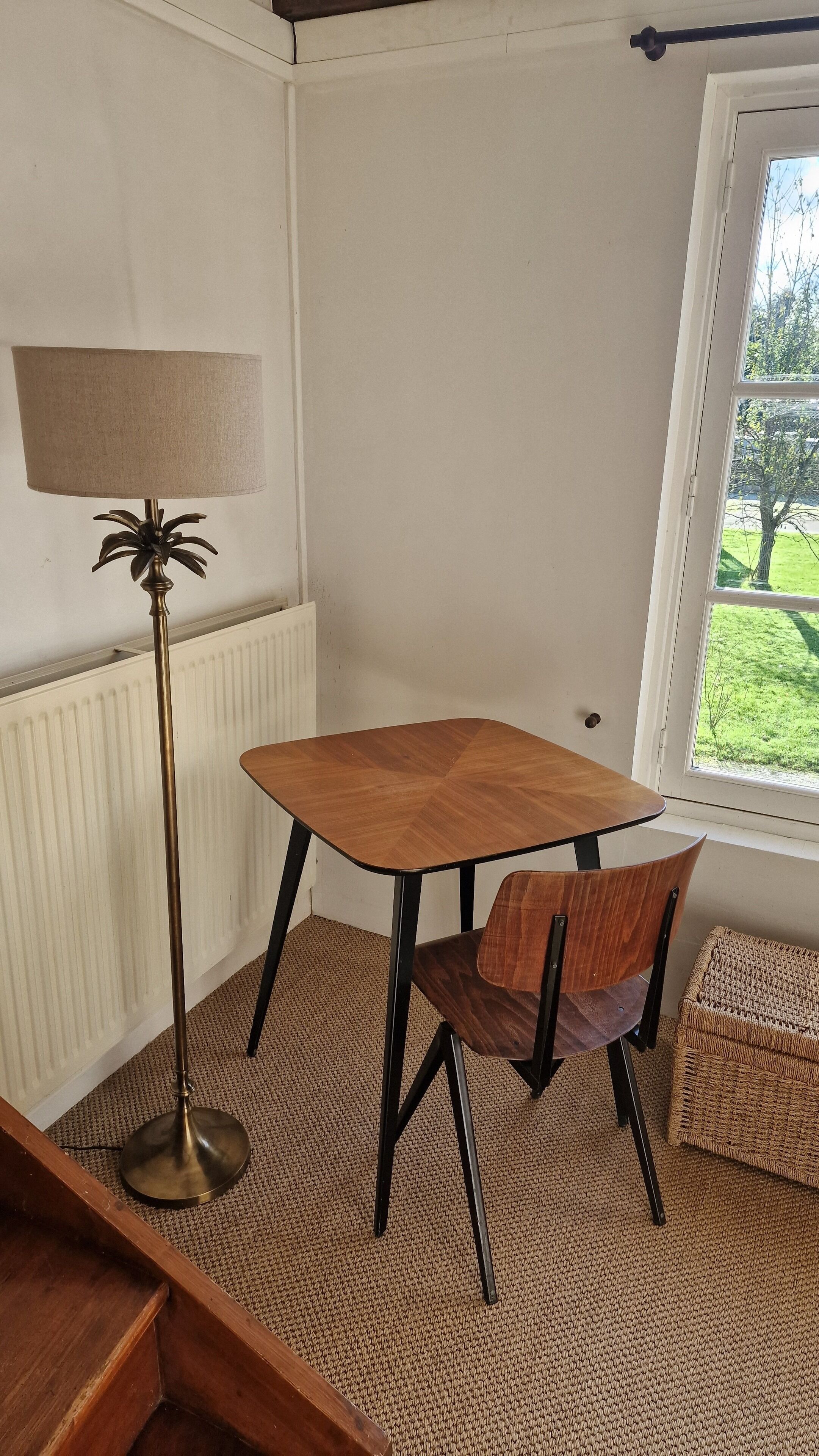 Een hoekzaal met een houten tafel, een stoel, een vloerlamp en een raam met uitzicht op groen.