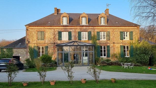 A large stone building with a symmetrical facade, multiple windows with shutters, a central entrance with a glass door, and a small covered porch.