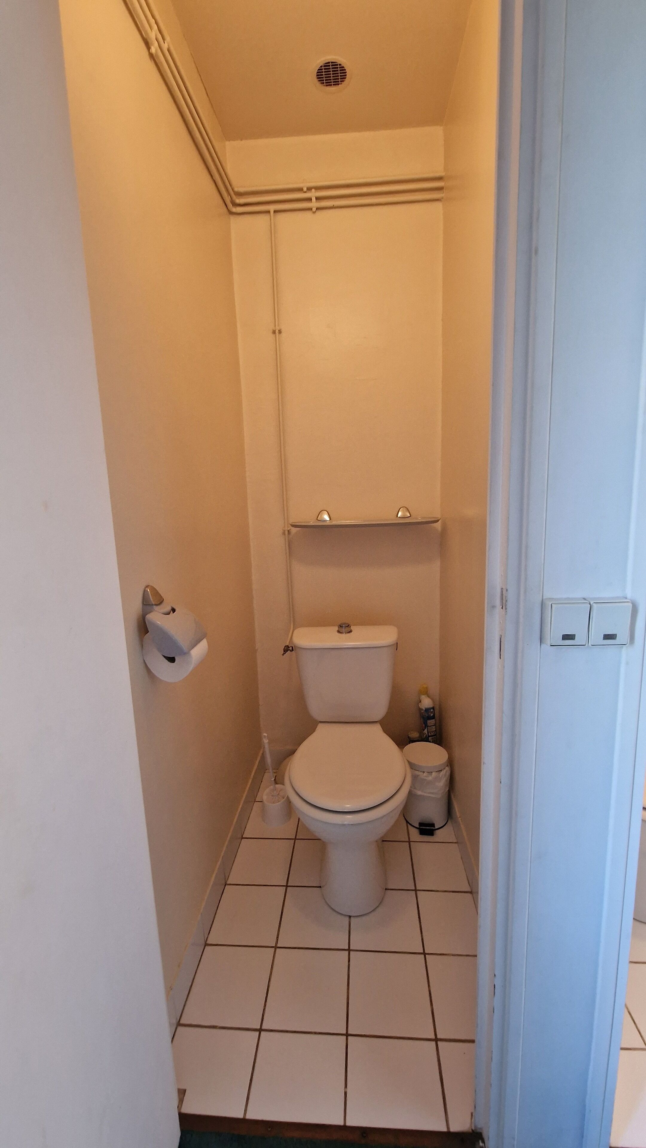 A small, narrow bathroom with a toilet, a shelf above the toilet, and a trash can.