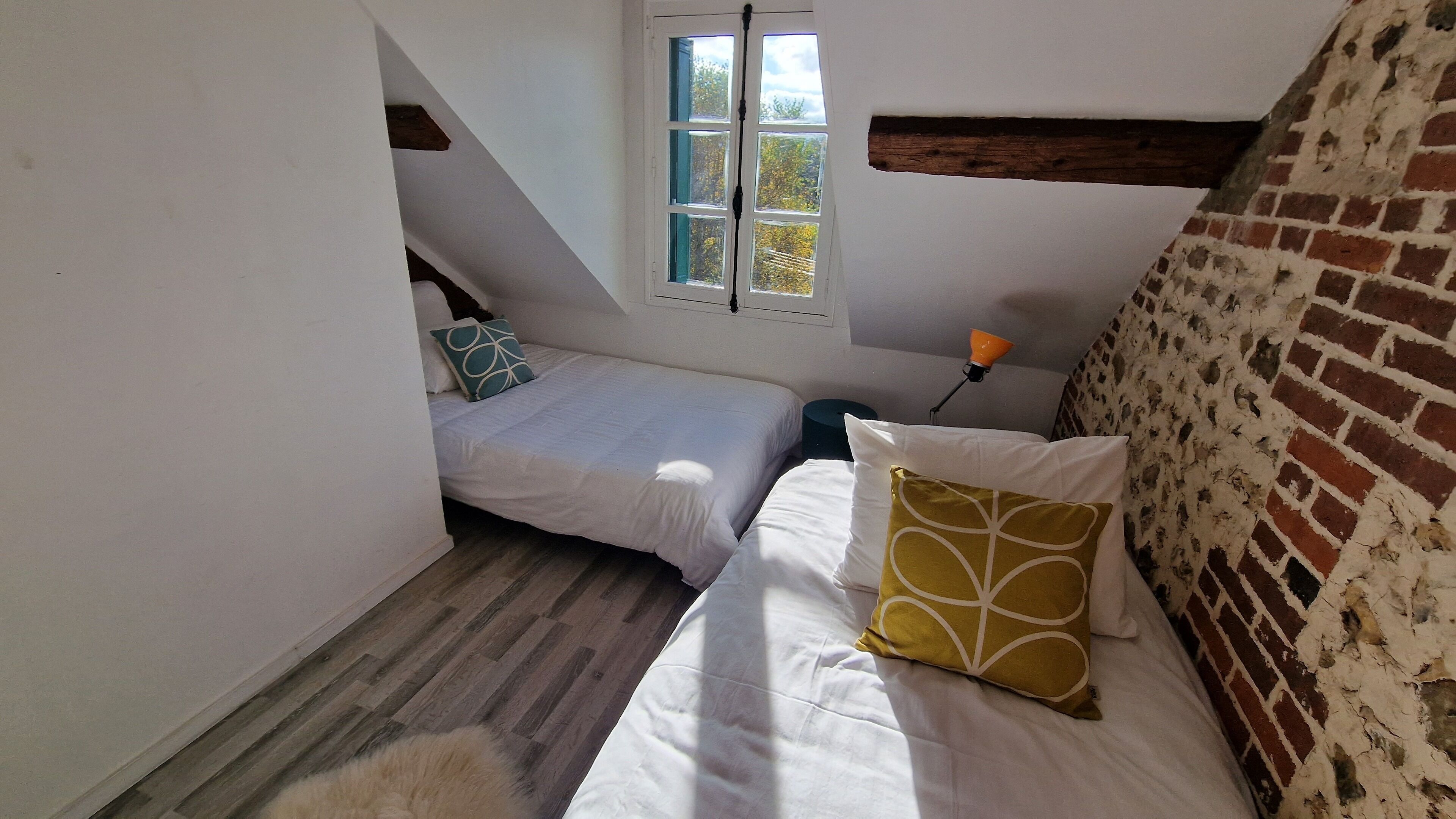 A small, attic-style bedroom with two beds, a window with a view, and a brick wall.
