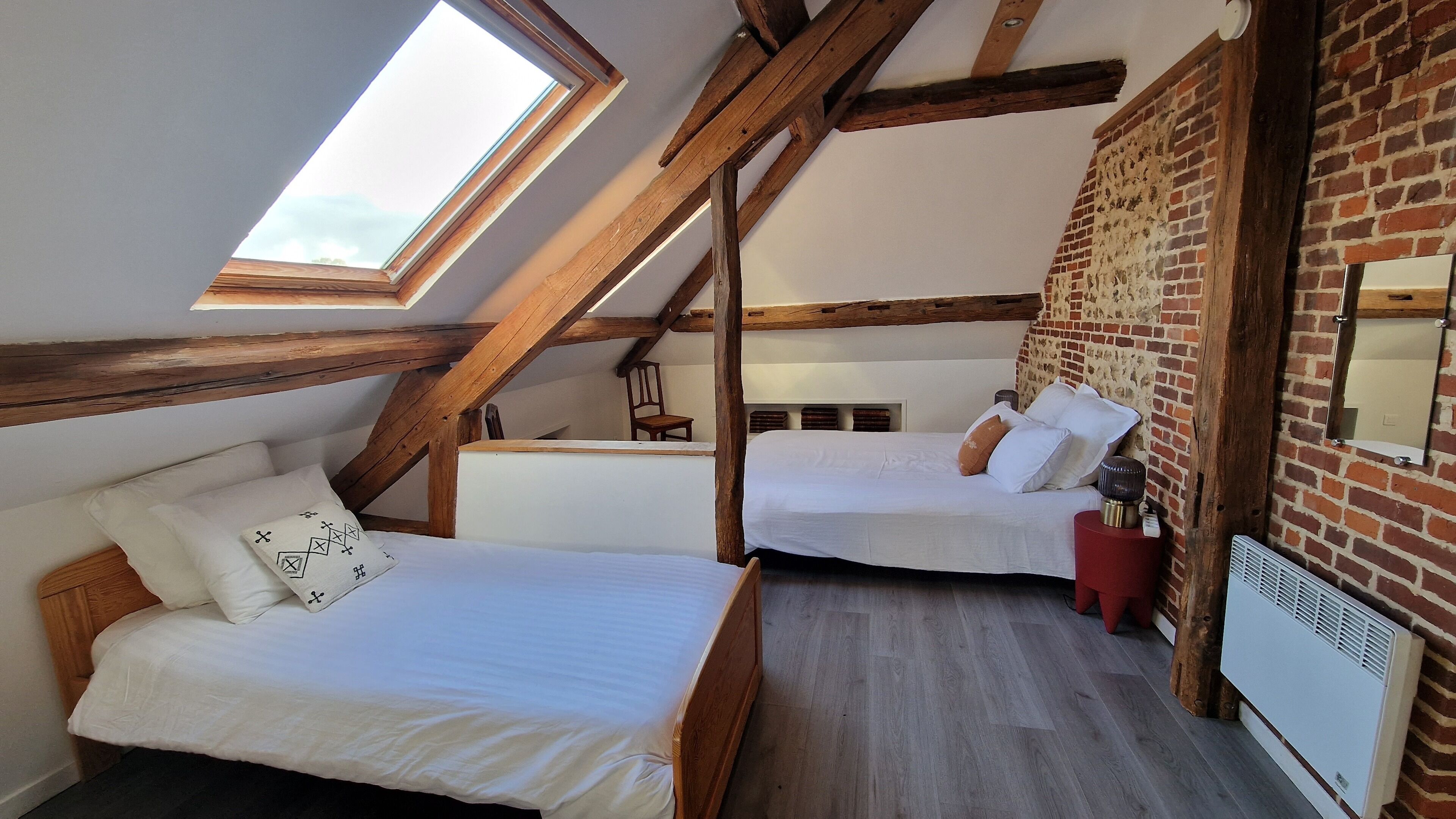 Une chambre aménagée en mezzanine avec deux lits, une lucarne, des poutres apparentes, un mur en briques et un radiateur.