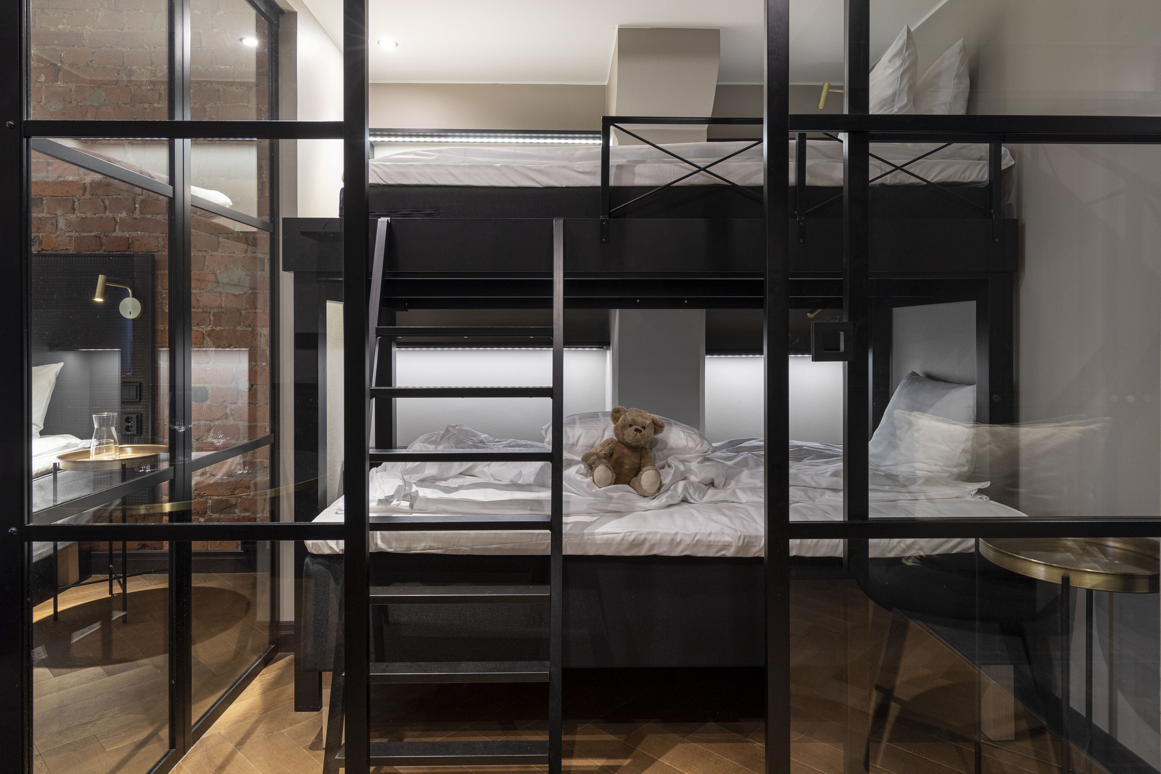 Une chambre avec des lits superposés séparés par une cloison en verre, un ours en peluche sur le lit du bas et une petite table avec une lampe.