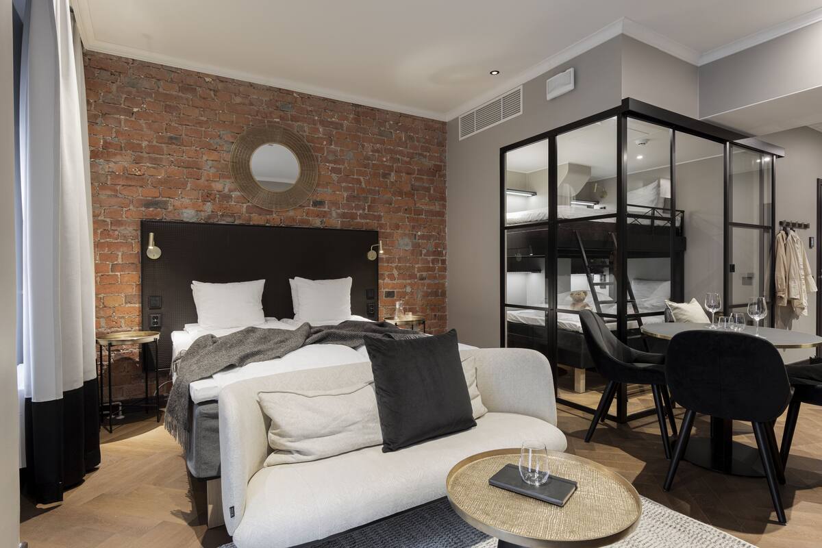 A modern hotel room with a brick wall, a bed with white linens, a sofa, a dining table with chairs, and a mirrored closet.
