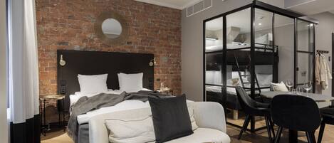 Una habitación de hotel moderna con pared de ladrillo, cama con ropa blanca, sofá, mesa de comedor con sillas y armario con espejo.